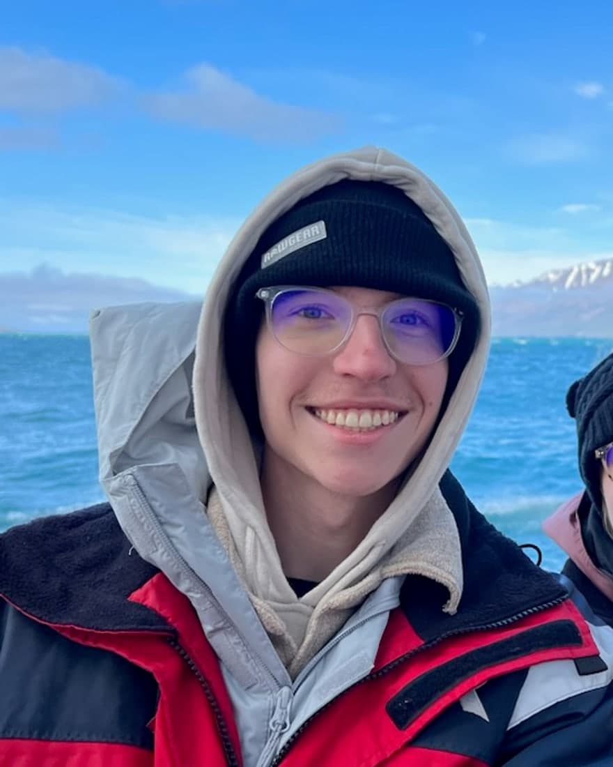 Ian Riley, smiling in a parka on a boat with a snow-capped coastline behind